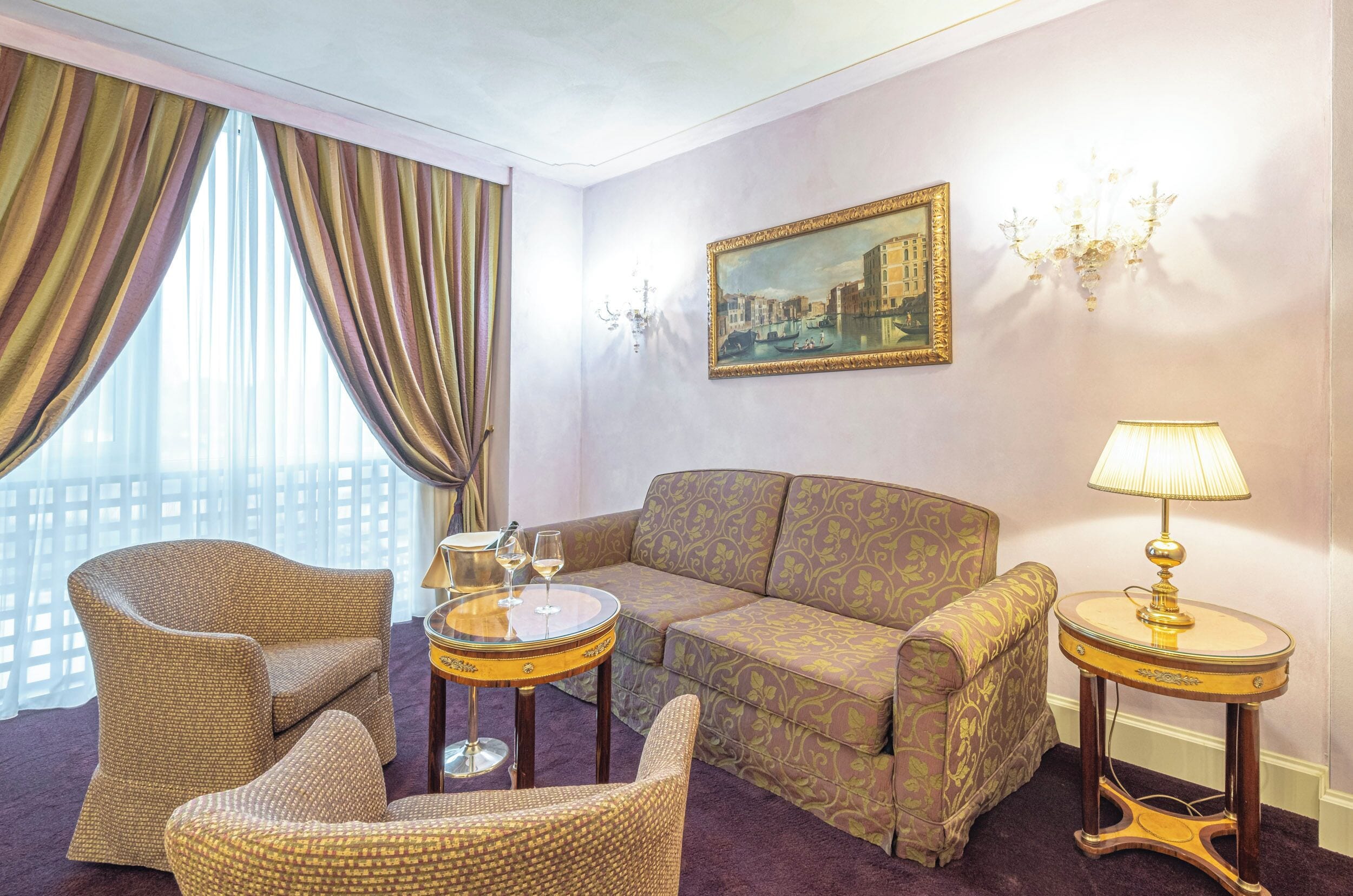 Quarto de hotel com um sofá estampado, duas poltronas, uma pequena mesa redonda com uma luminária e um quadro na parede.