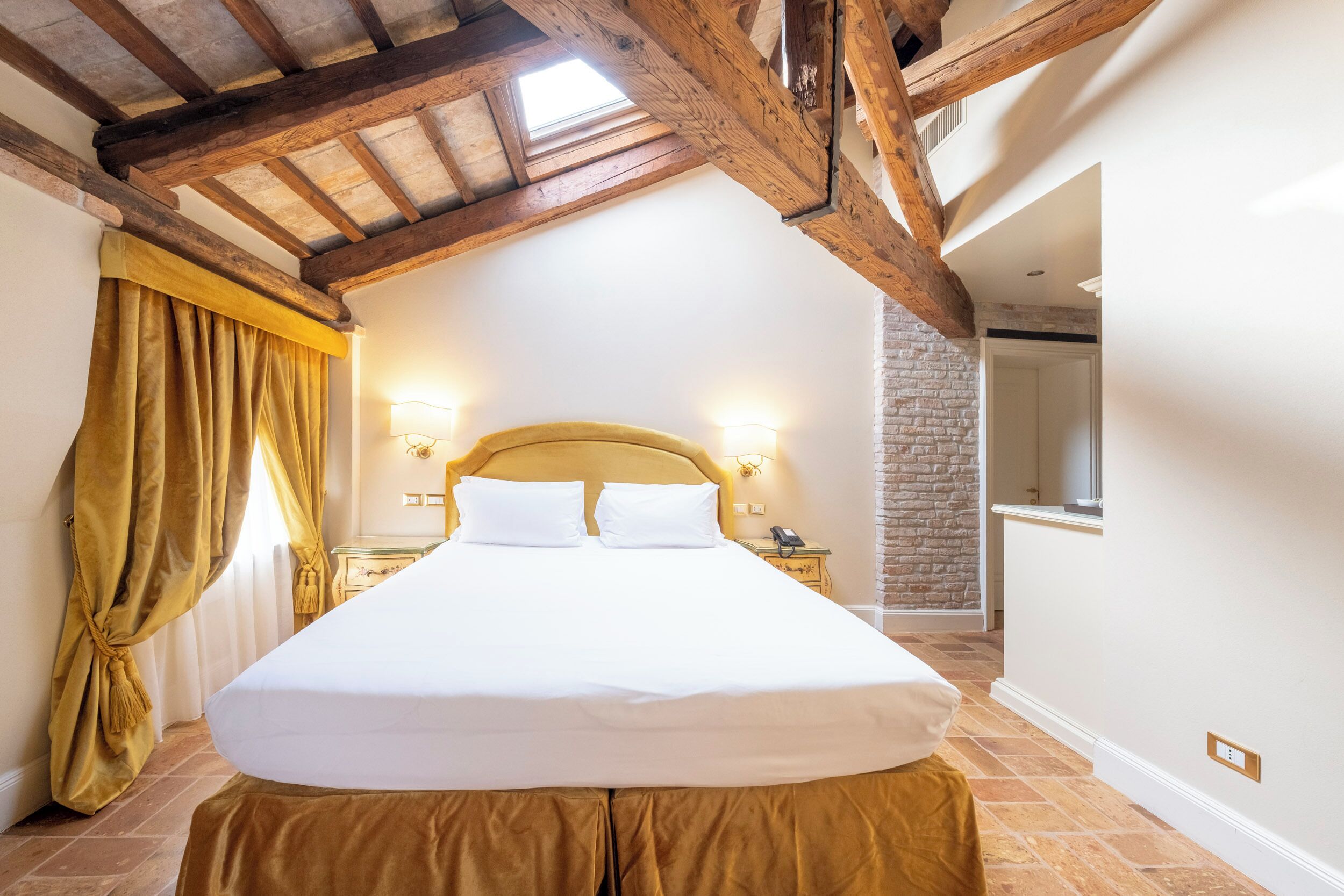 A bedroom with a large bed, wooden beams, a stone wall, and a window.