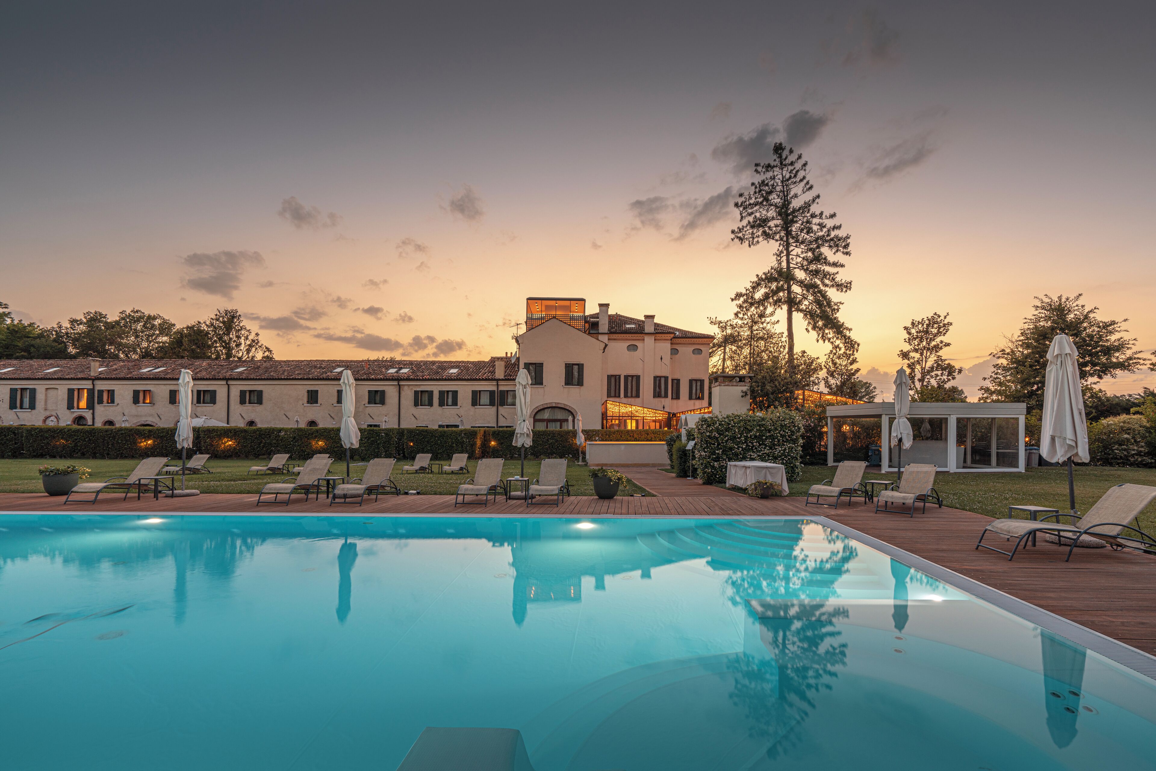 Piscina con sillas de descanso y sombrillas, rodeada por un jardín ajardinado y un edificio con múltiples ventanas.