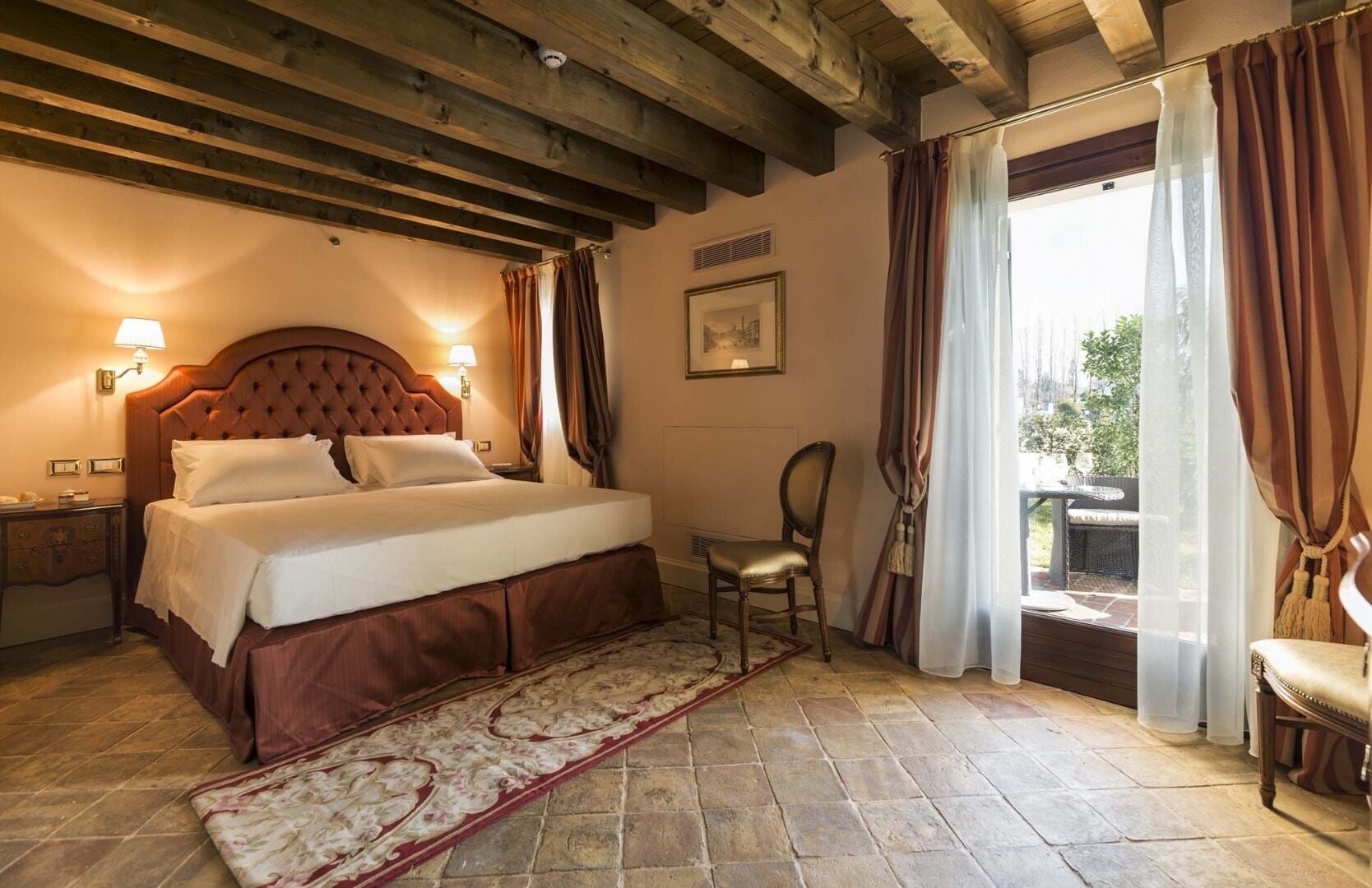 Un dormitorio con una cama grande, cabecera de madera, mesita de noche, una silla y vistas al exterior a través de una ventana con cortinas.
