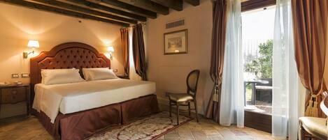 Un dormitorio con una cama grande, cabecera de madera, mesita de noche, una silla y vistas al exterior a través de una ventana con cortinas.