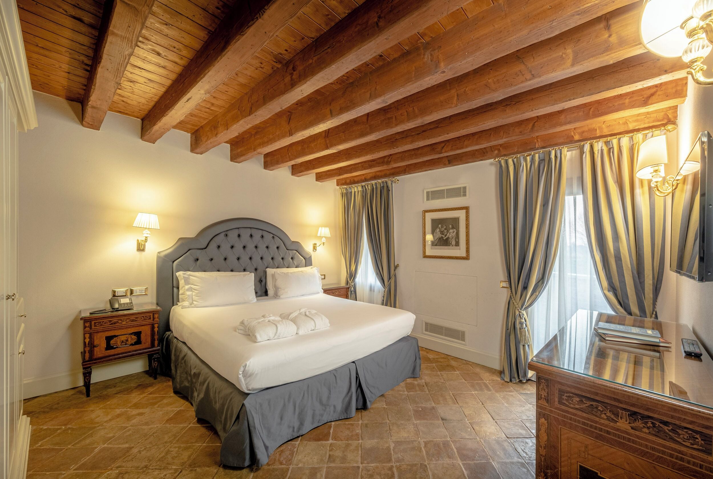 Une chambre à coucher avec un grand lit, des poutres apparentes en bois au plafond, une table de chevet, un bureau et une lampe.