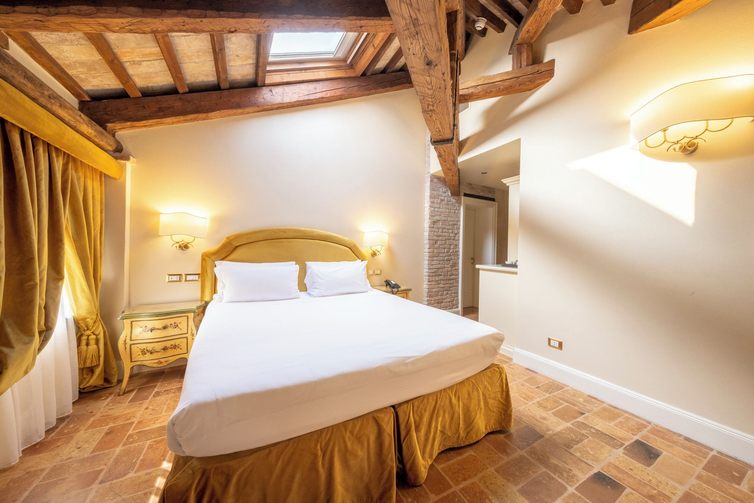 A bedroom with a large bed, wooden beams, a skylight, and a stone wall.