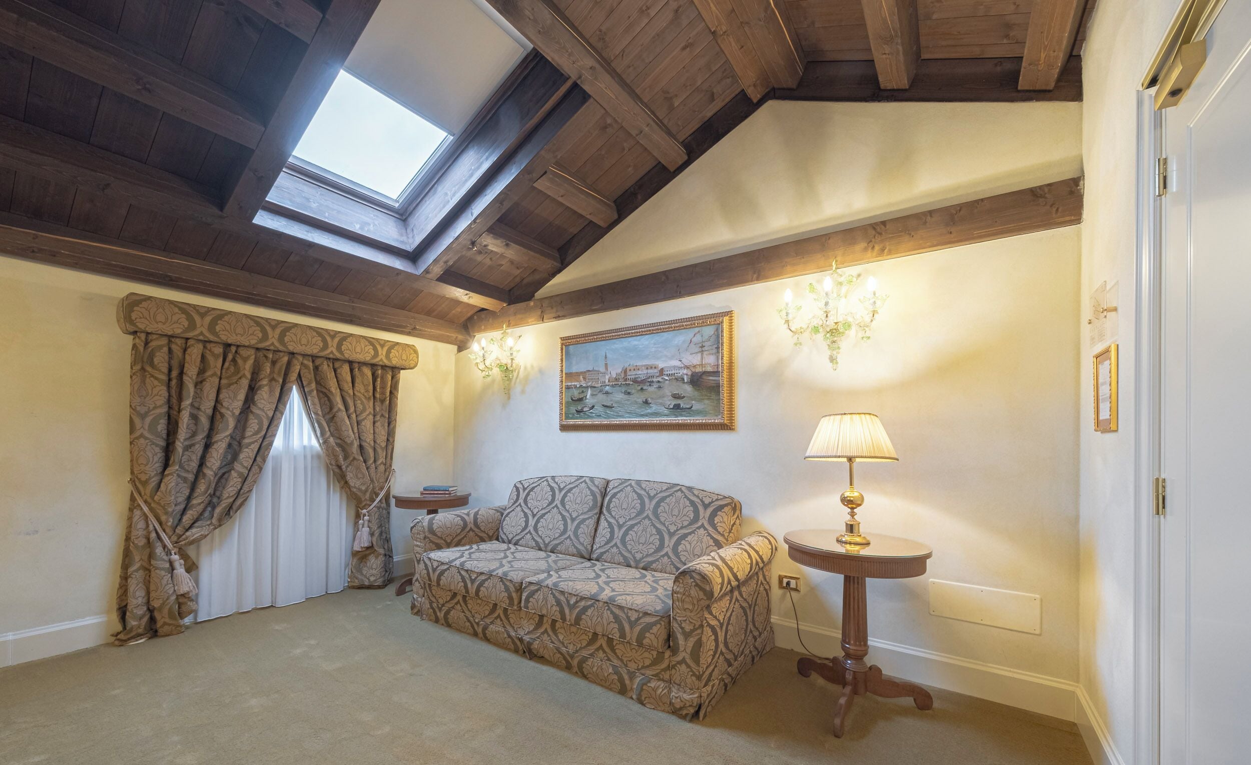 A cozy room with a sloped ceiling, a patterned sofa, a small round table with a lamp, and a painting of a cityscape.