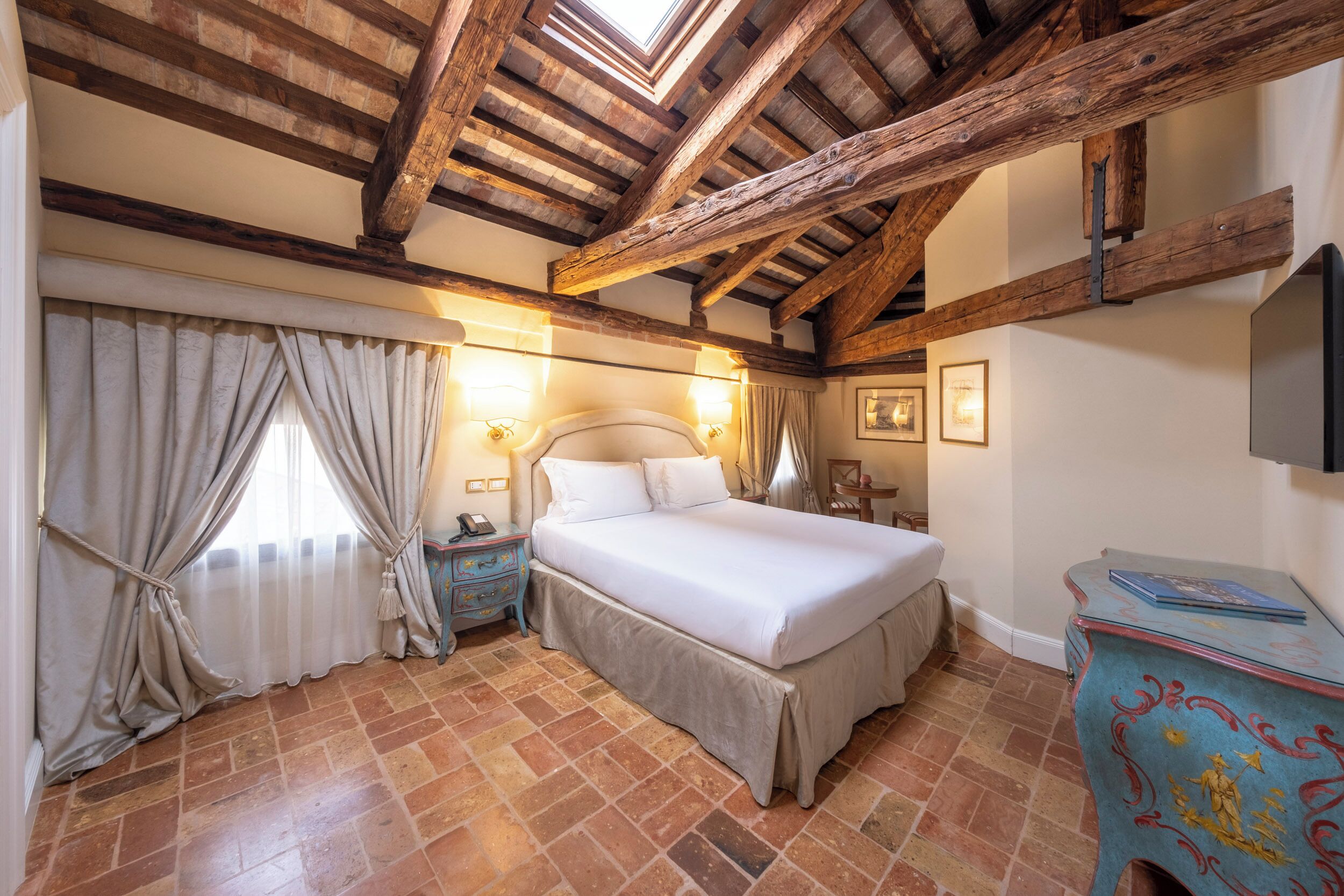Un dormitorio con una cama grande, vigas de madera, una mesita de noche, un aparador y una ventana con cortinas.