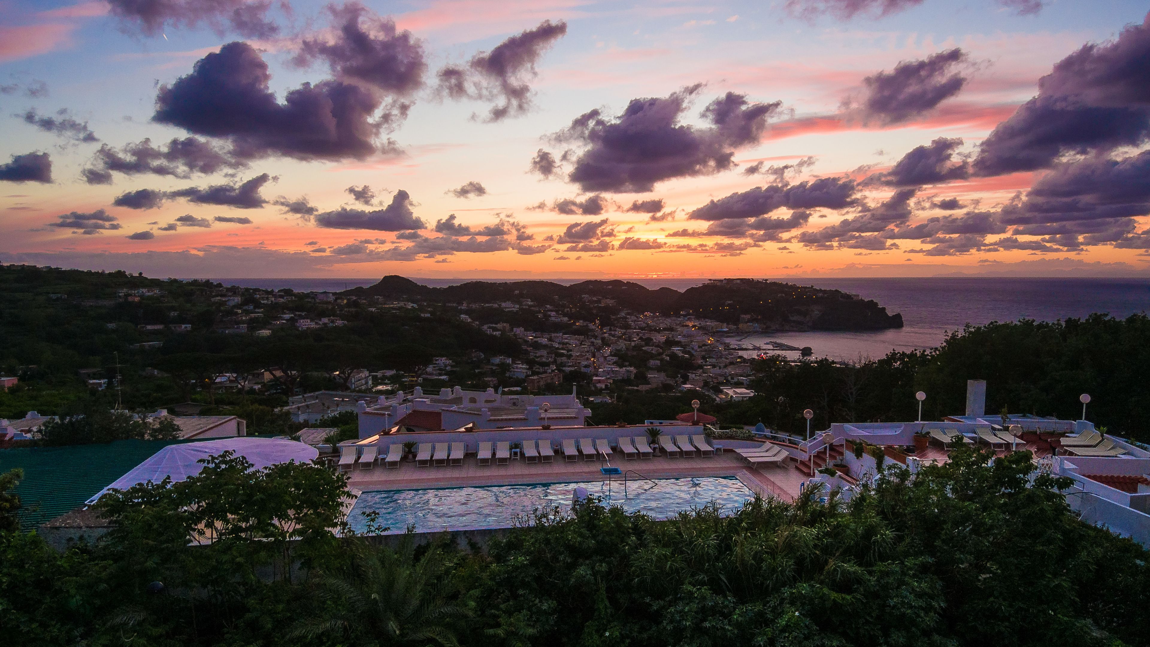 Un resort con piscina, sillones de descanso y vistas al mar y al pueblo al atardecer.