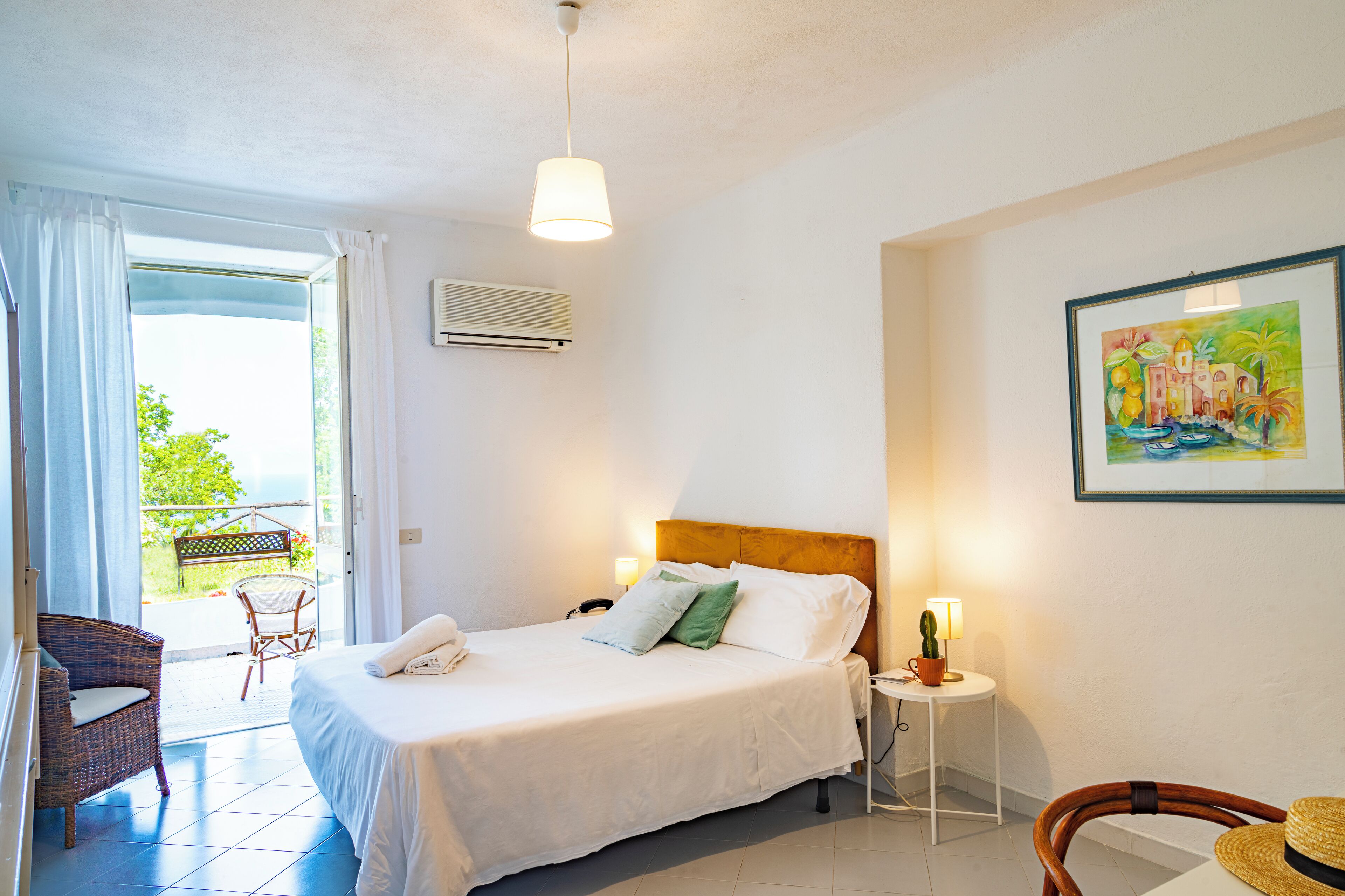 A hotel room with a bed, a chair, a small table with a lamp, and a painting on the wall.