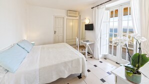 Un dormitorio con cama, escritorio, silla, una orquídea en maceta y vistas al mar a través de amplios ventanales.