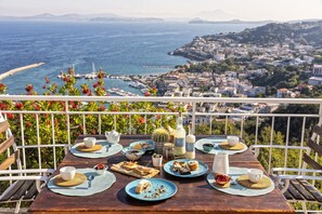A wooden table set for a meal with a view of the sea and a coastal town. - Relais Paradise Villa Janto' (Casamicciola Terme)