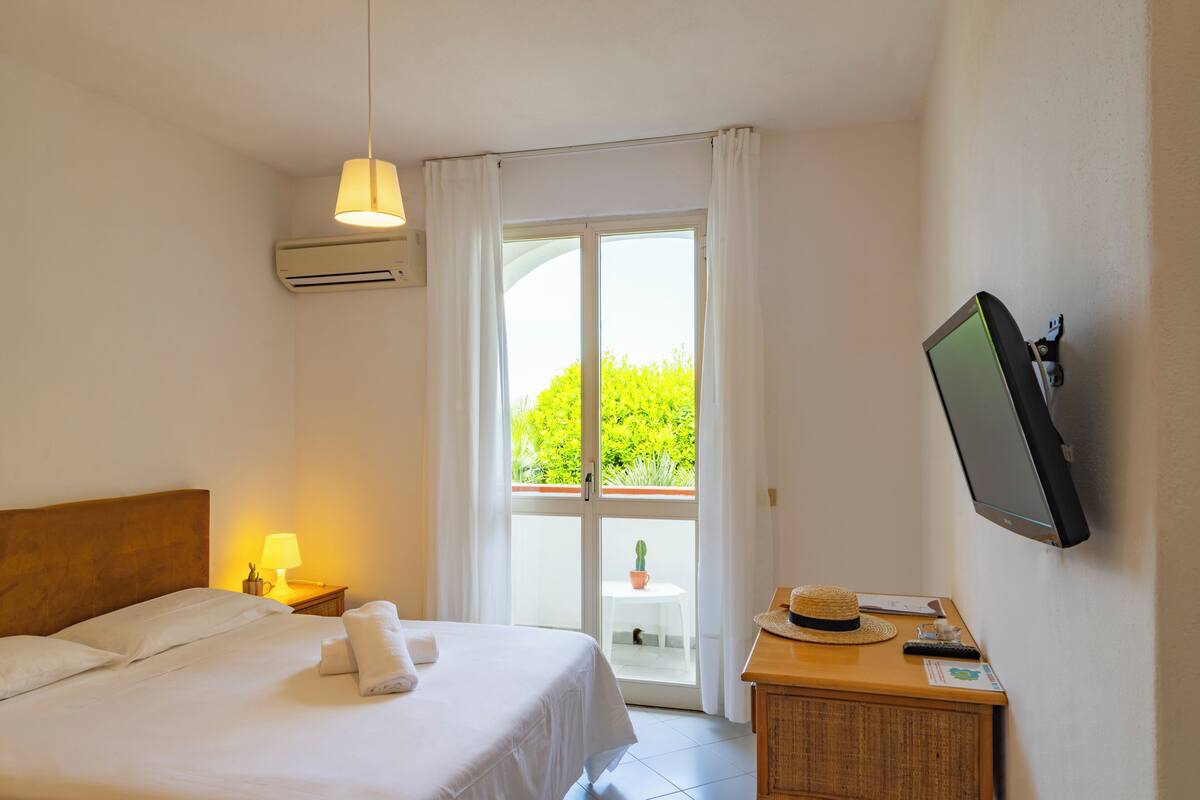 A hotel room with a bed, a desk with a television, a lamp, and a view of greenery outside.