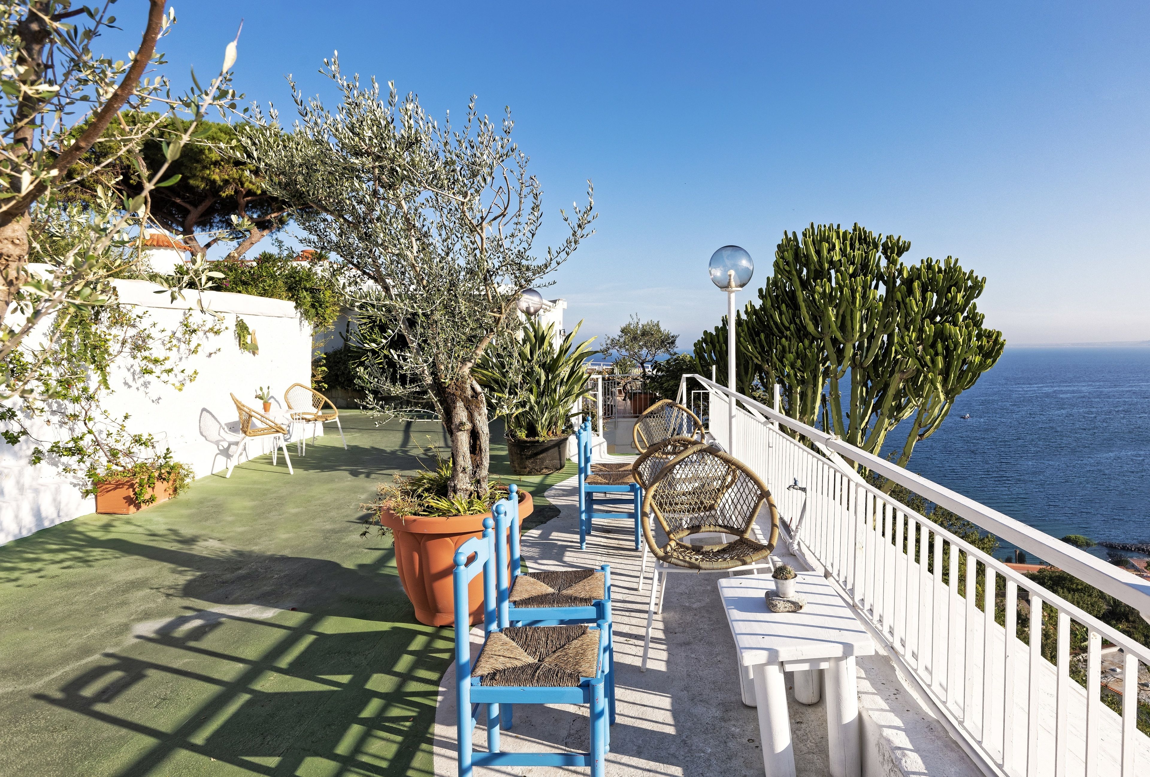 Terraza costera con sillas azules, plantas en macetas y una baranda blanca con vista al mar.