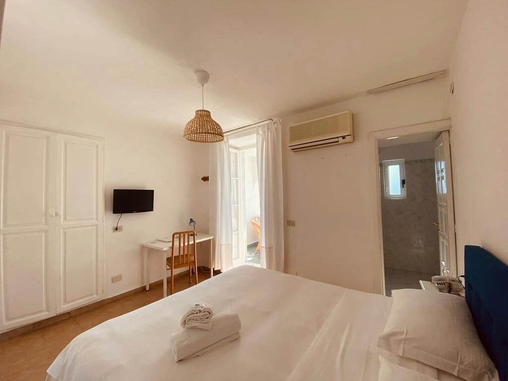 A hotel room with a bed, a desk, a chair, a television, and an air conditioning unit.