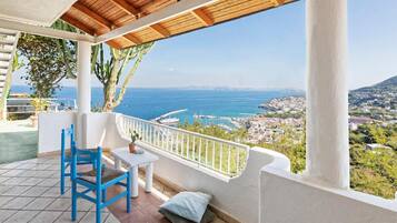 Un balcón con techo de madera, barandilla blanca, una silla azul, una mesita y vistas al mar y a un pueblo.