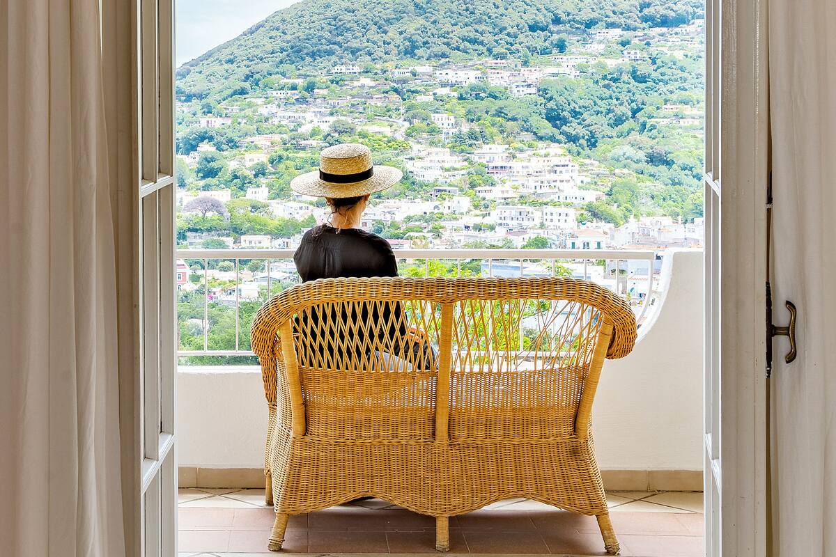 A wicker chair facing a balcony with a view of a hillside town.