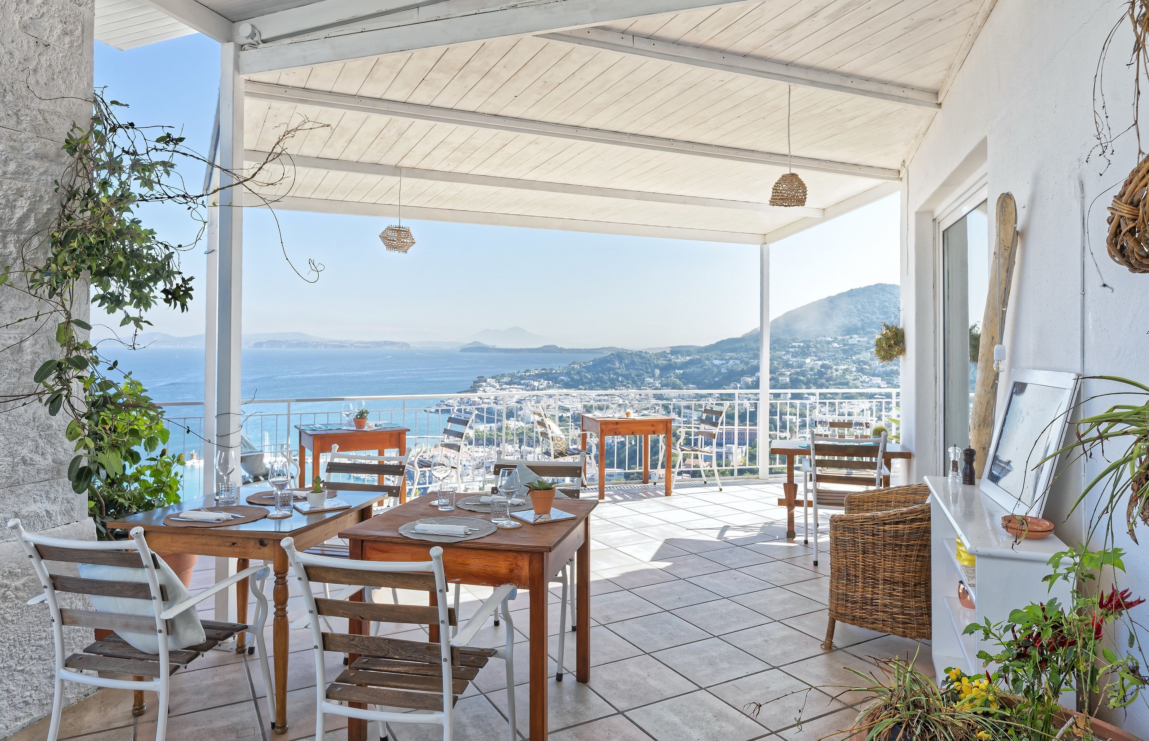 Um terraço coberto com uma mesa posta para refeições, com vista para uma vila costeira e o mar.