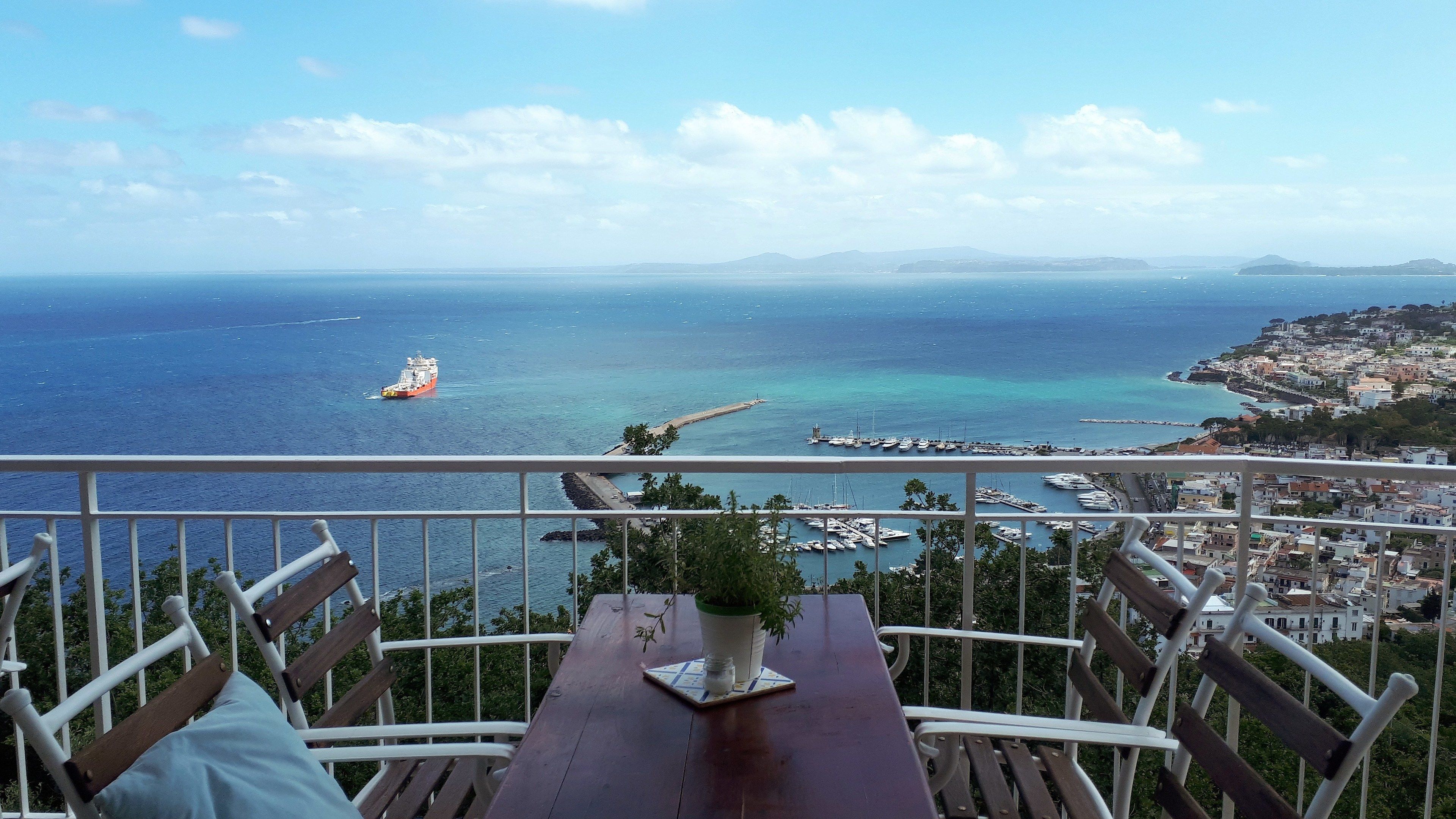 Ein Balkon mit Holztisch, einem Topfpflanzen und Blick auf das Meer mit Booten und einer fernen Küste.