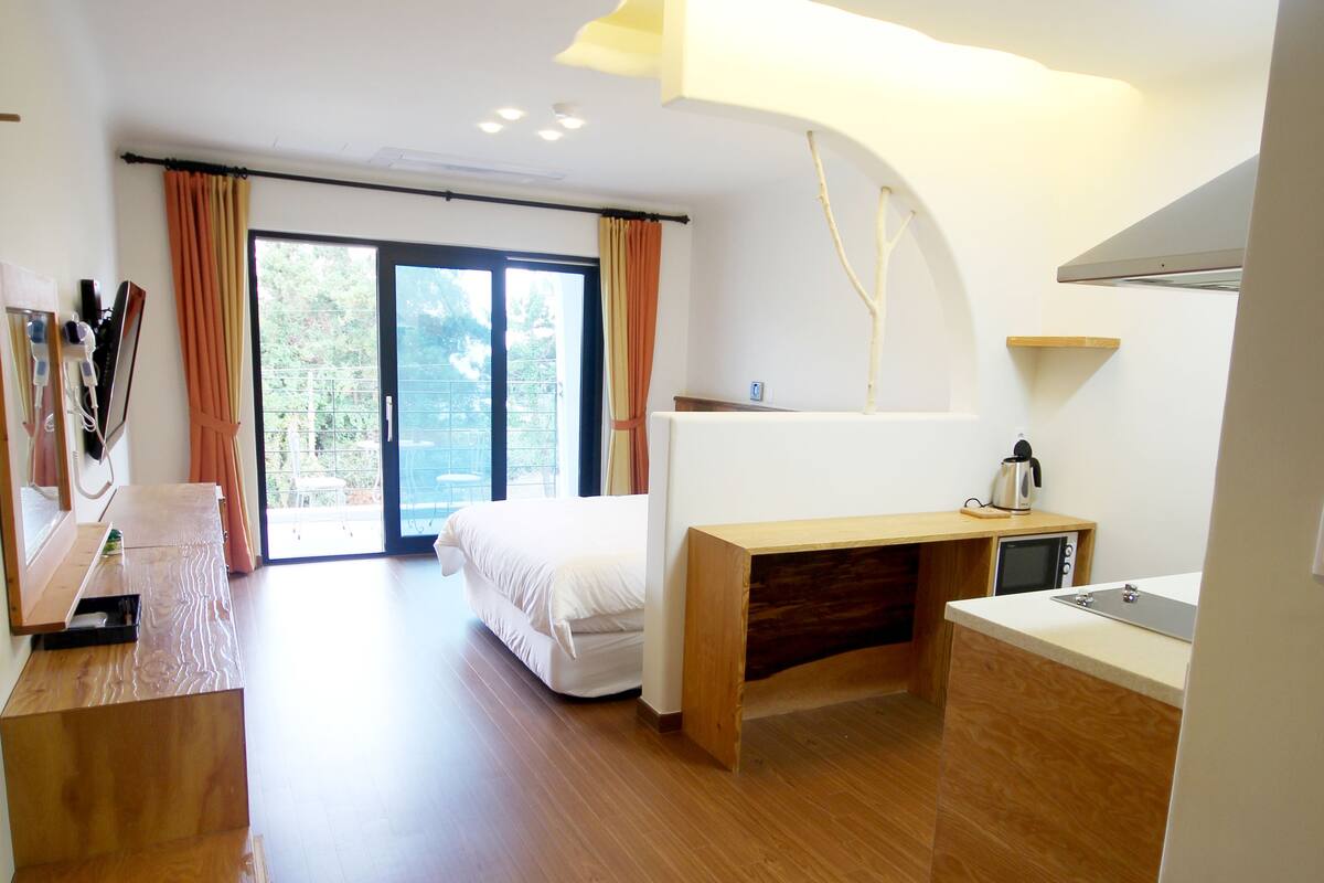 A modern hotel room with a wooden floor, a bed, a desk, a small kitchenette, and a sliding glass door leading to an outdoor area.