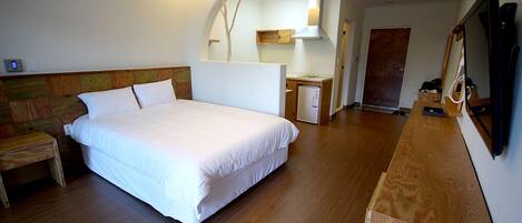 A bedroom with a large bed, a wooden headboard, a small bedside table, a nightstand, a wooden dresser, a mirror, a television, and a kitchenette with a stove and sink.