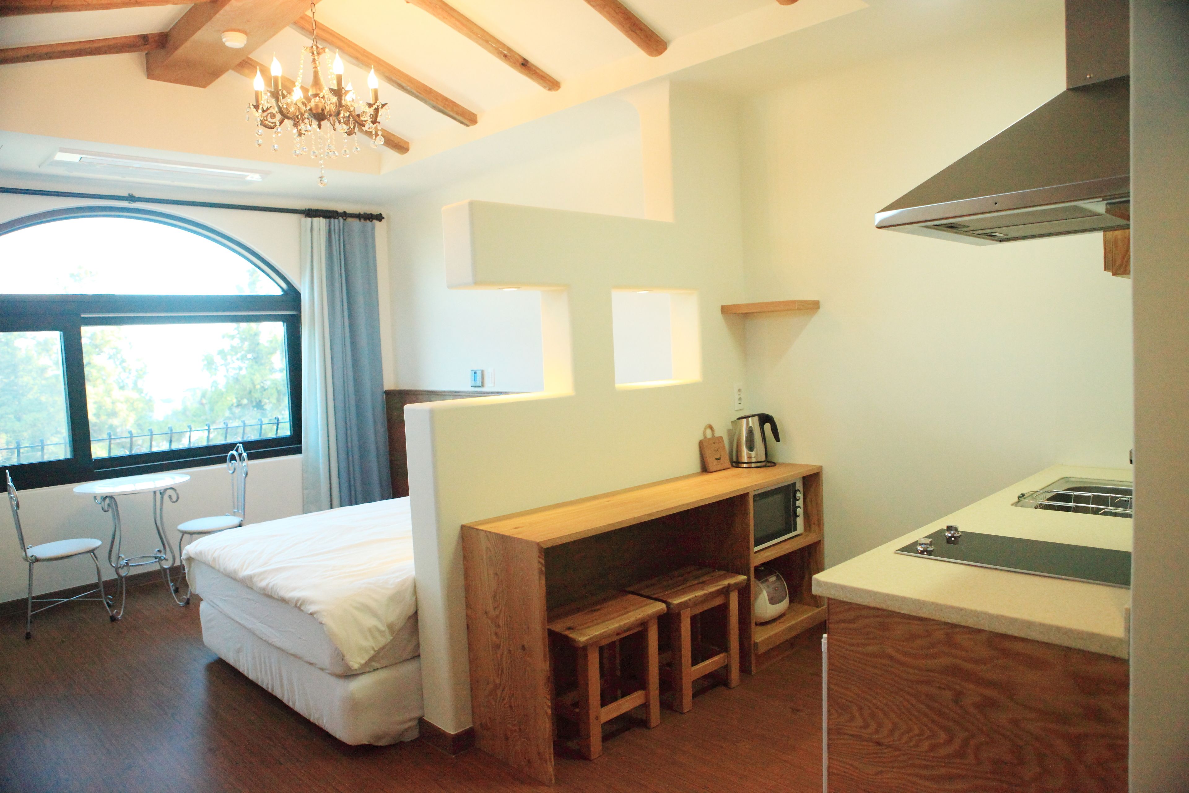 A bedroom with a bed, a small table, two chairs, a window with curtains, a wooden bench, a TV, and a kitchenette.