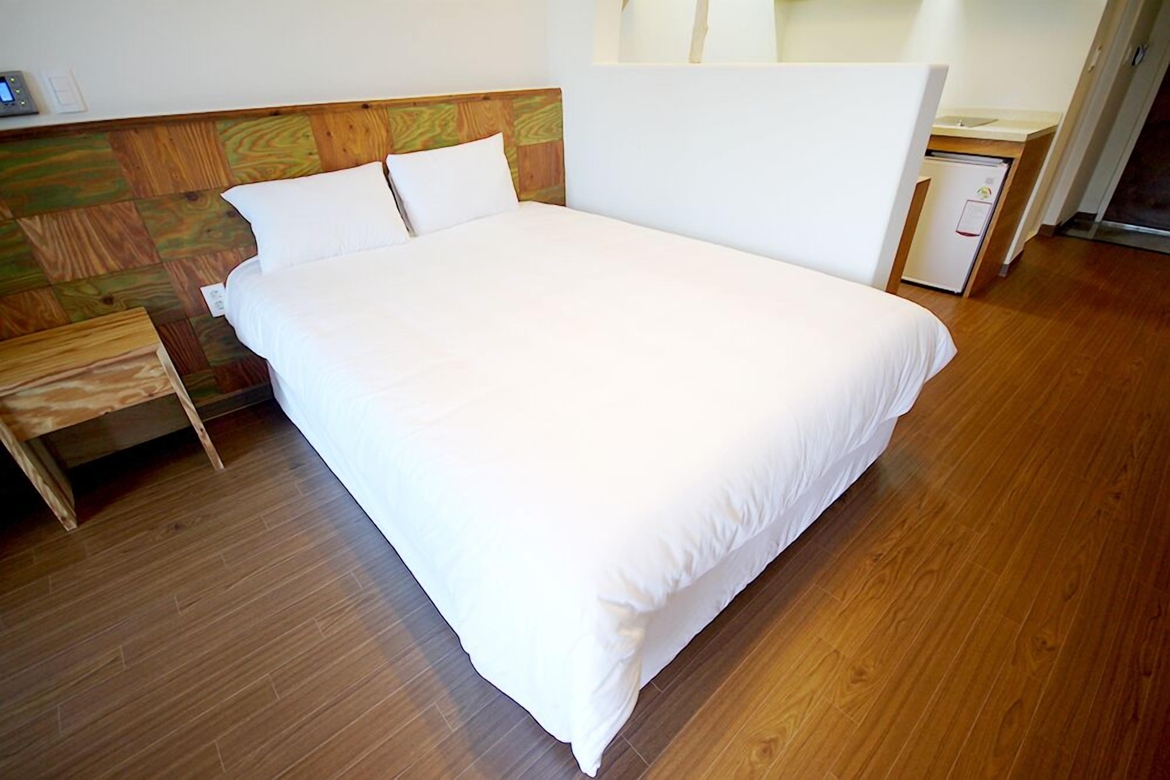 A neatly made bed with white linens in a room with wooden flooring and a wooden headboard.
