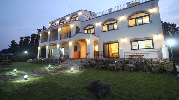 A two-story house with multiple balconies, large windows, and a well-lit exterior. - Nomad Pension in Jeju (Seogwipo)