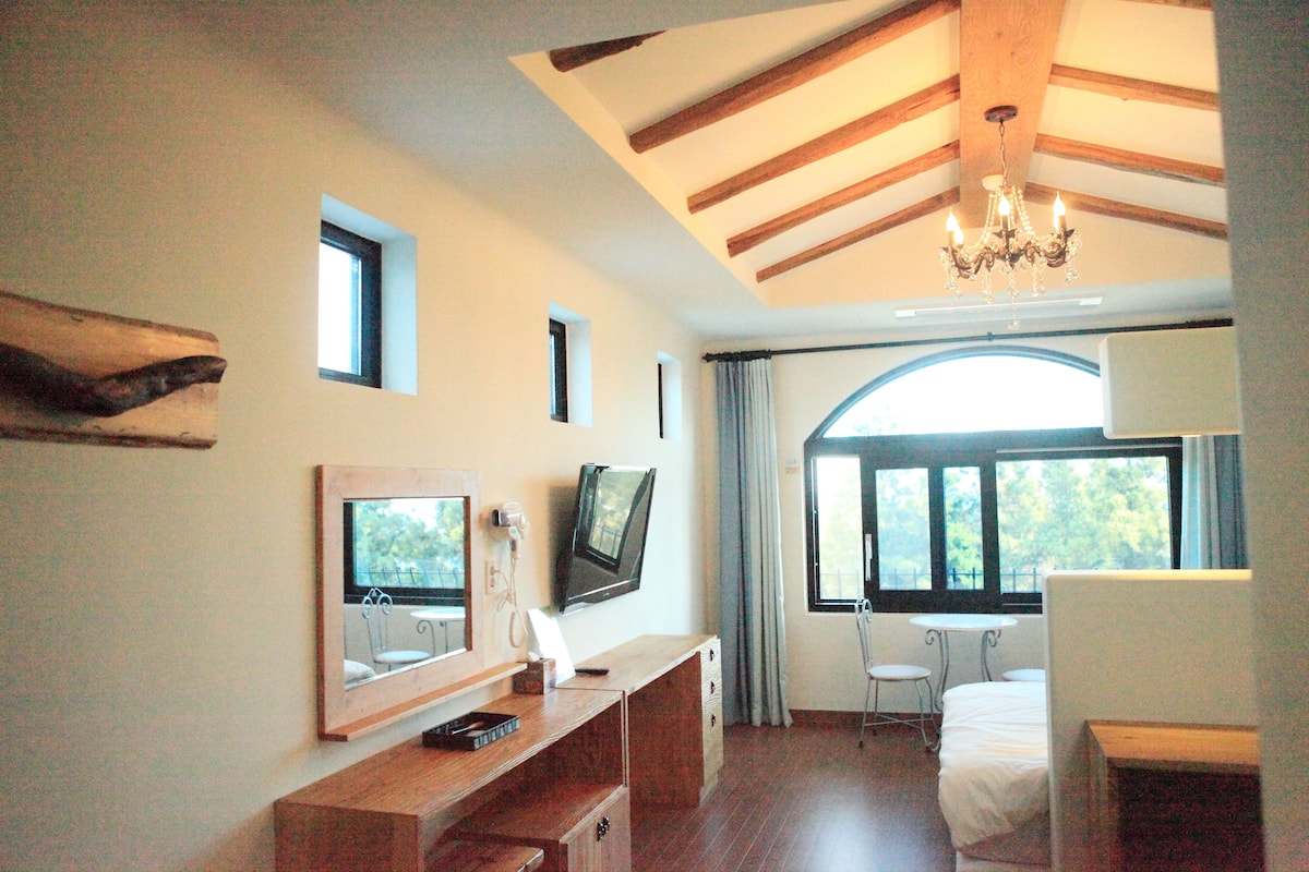 A bedroom with a wooden ceiling, a chandelier, a bed, a desk with a mirror, a television, and a window with curtains.