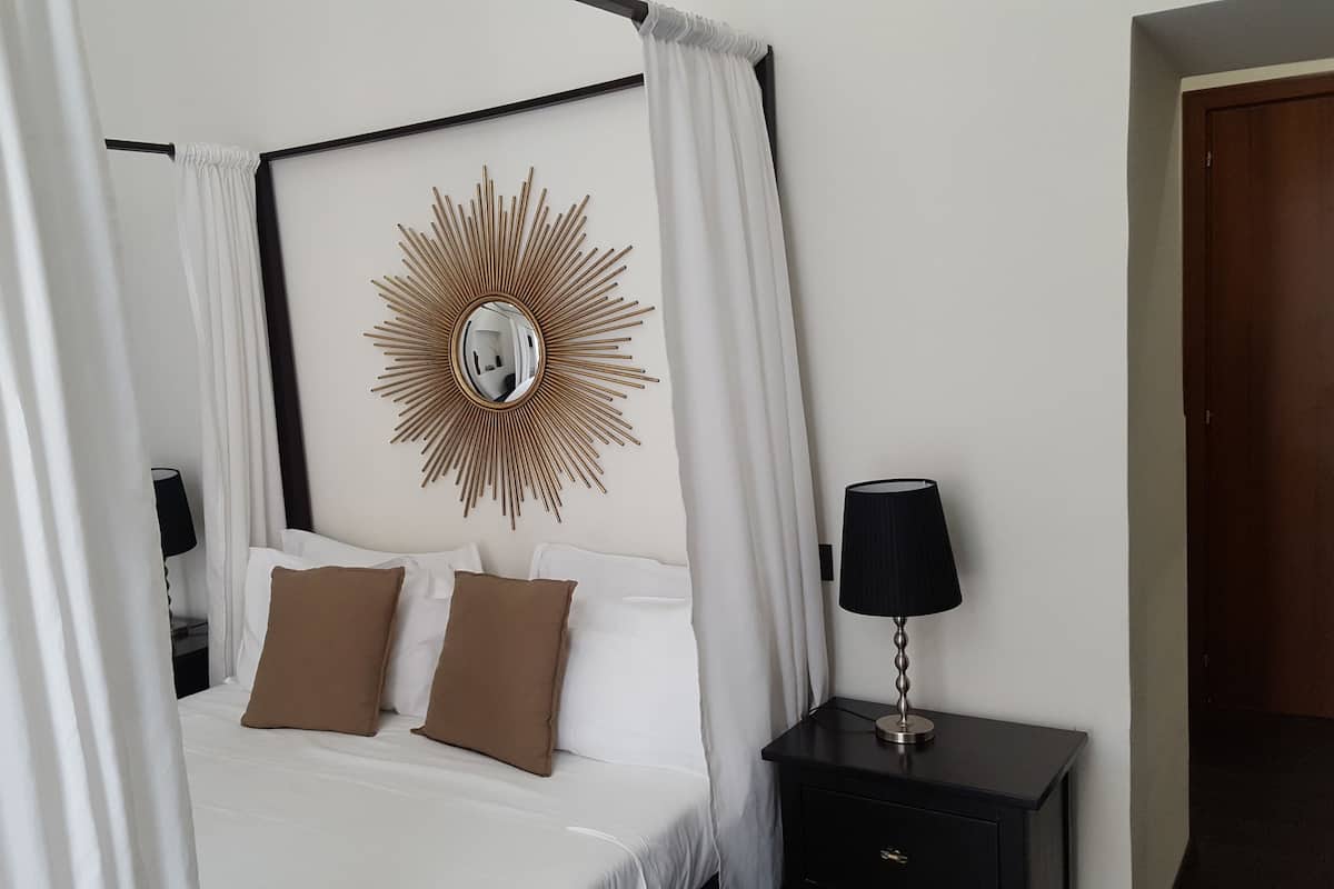 A four-poster bed with white and brown bedding, a sunburst mirror on the wall, and a black nightstand with a lamp.