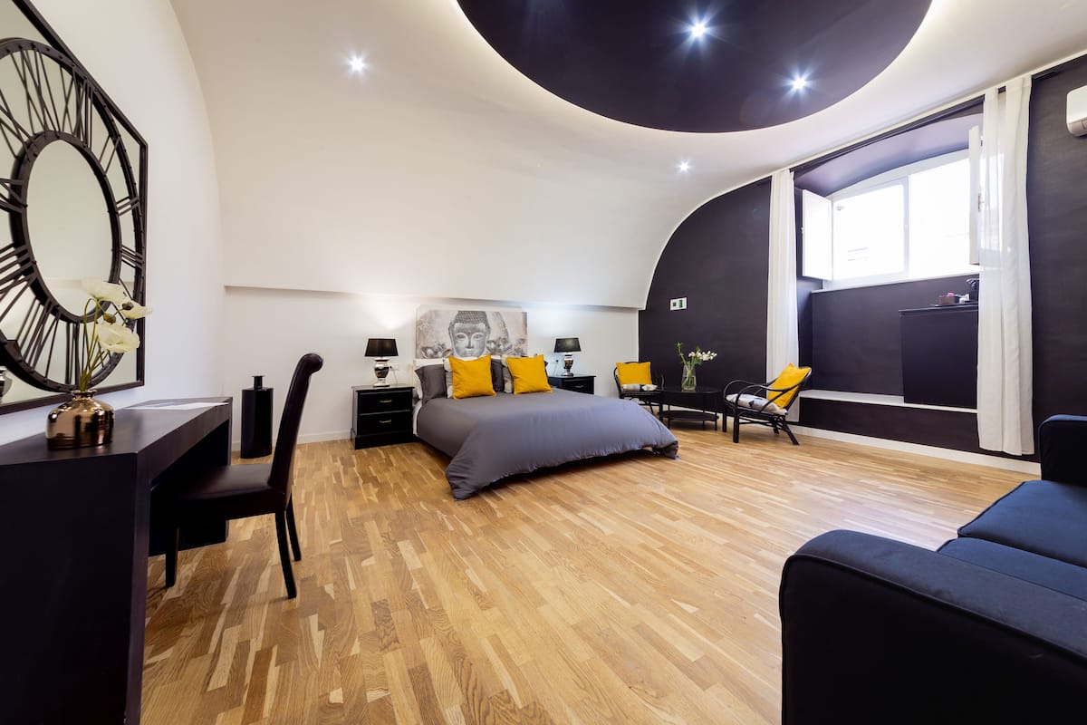 A modern bedroom with a large bed, a desk, and a chair. There is a window with curtains, a wall-mounted mirror, and a Buddha statue.
