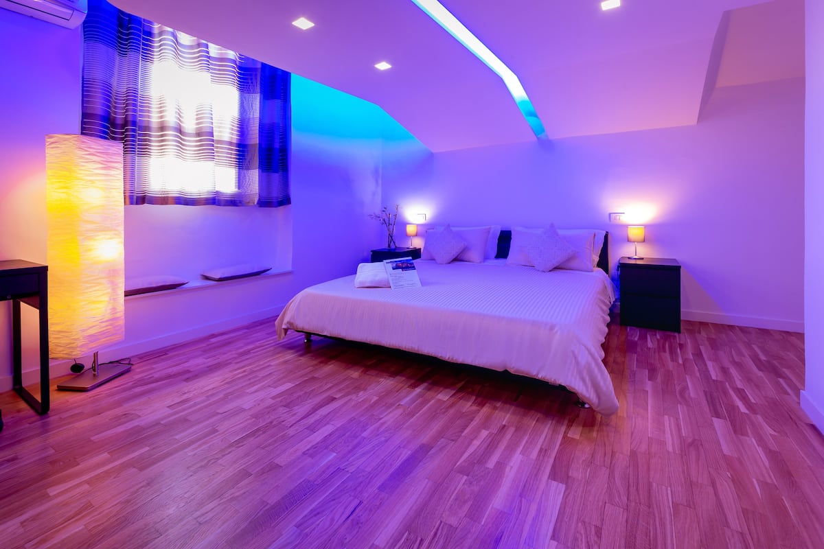 A modern bedroom with a large bed, wooden flooring, and ambient lighting.