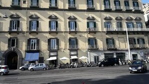 Un edificio de varios pisos con una fila de balcones, una entrada central con un arco de piedra y mobiliario exterior con sombrillas.