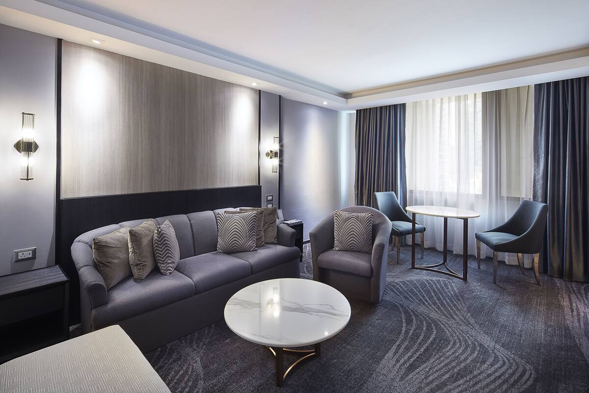 A modern hotel room with a grey sofa, two armchairs, a round coffee table, and a small side table.
