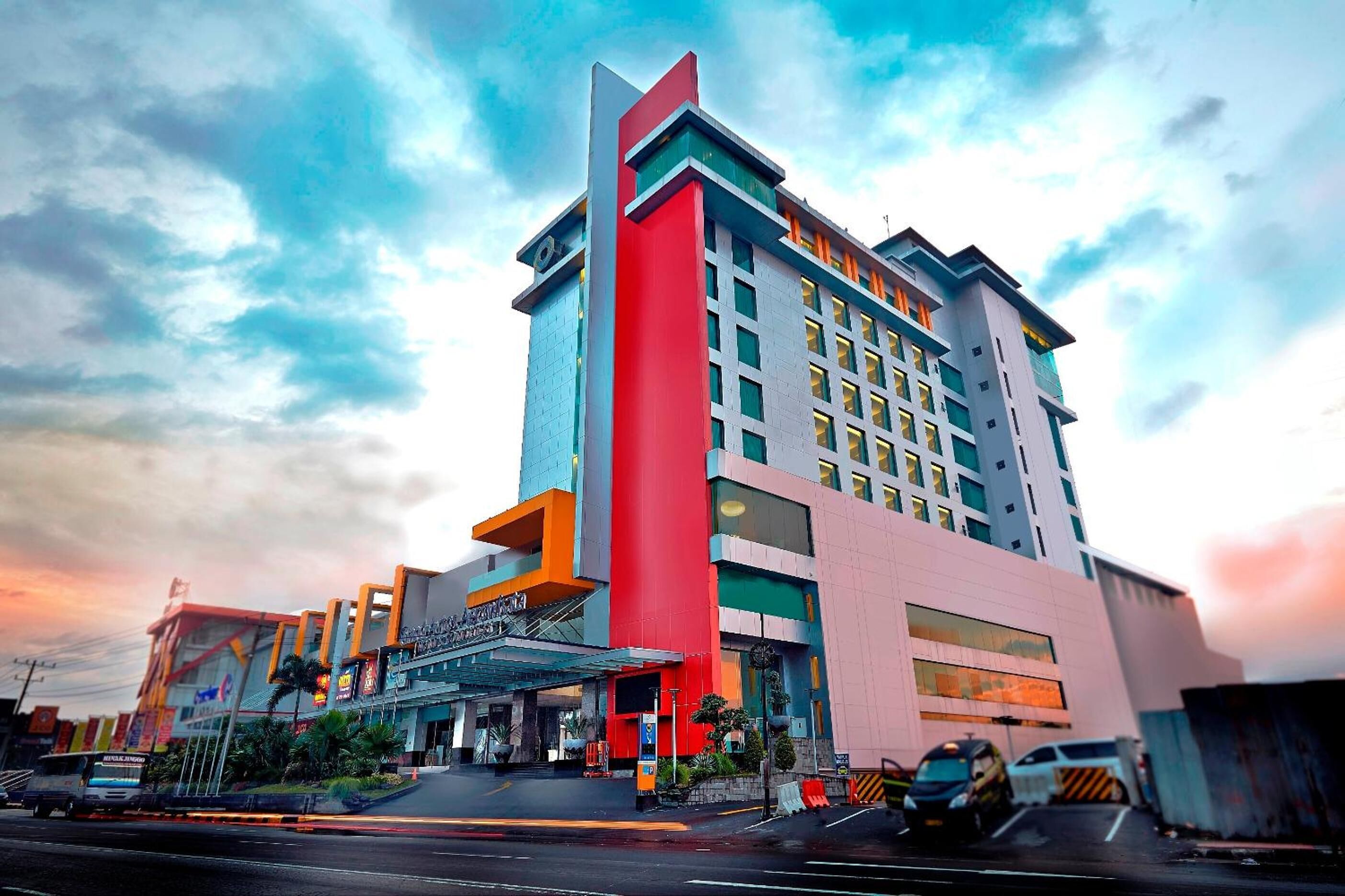 A multi-story hotel with a prominent red and blue facade, a covered entrance, and a parking area with vehicles.