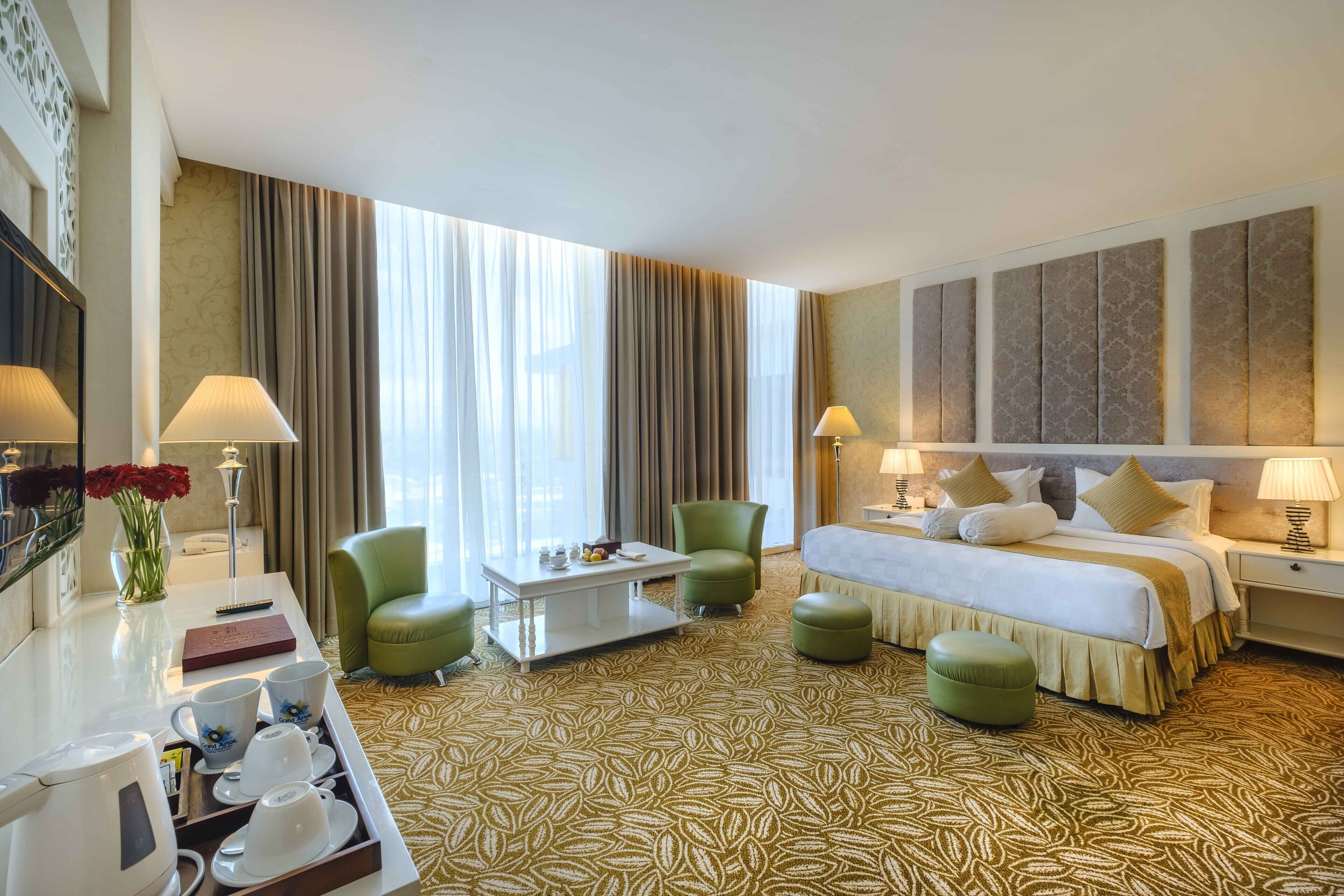 A hotel room with a bed, two green armchairs, a coffee table, a desk with a lamp, and a large window with curtains.