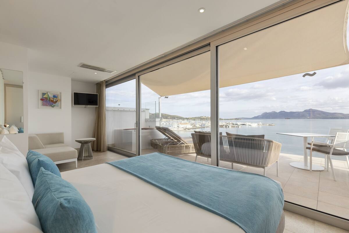 A modern hotel room with a large bed, a TV, a balcony with seating, and a view of the sea.