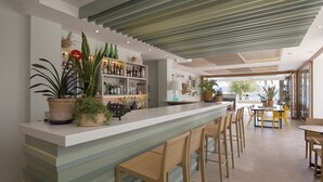 Un área de bar moderna con mostrador, taburetes y estanterías llenas de botellas. Hay plantas en macetas y vistas al exterior.