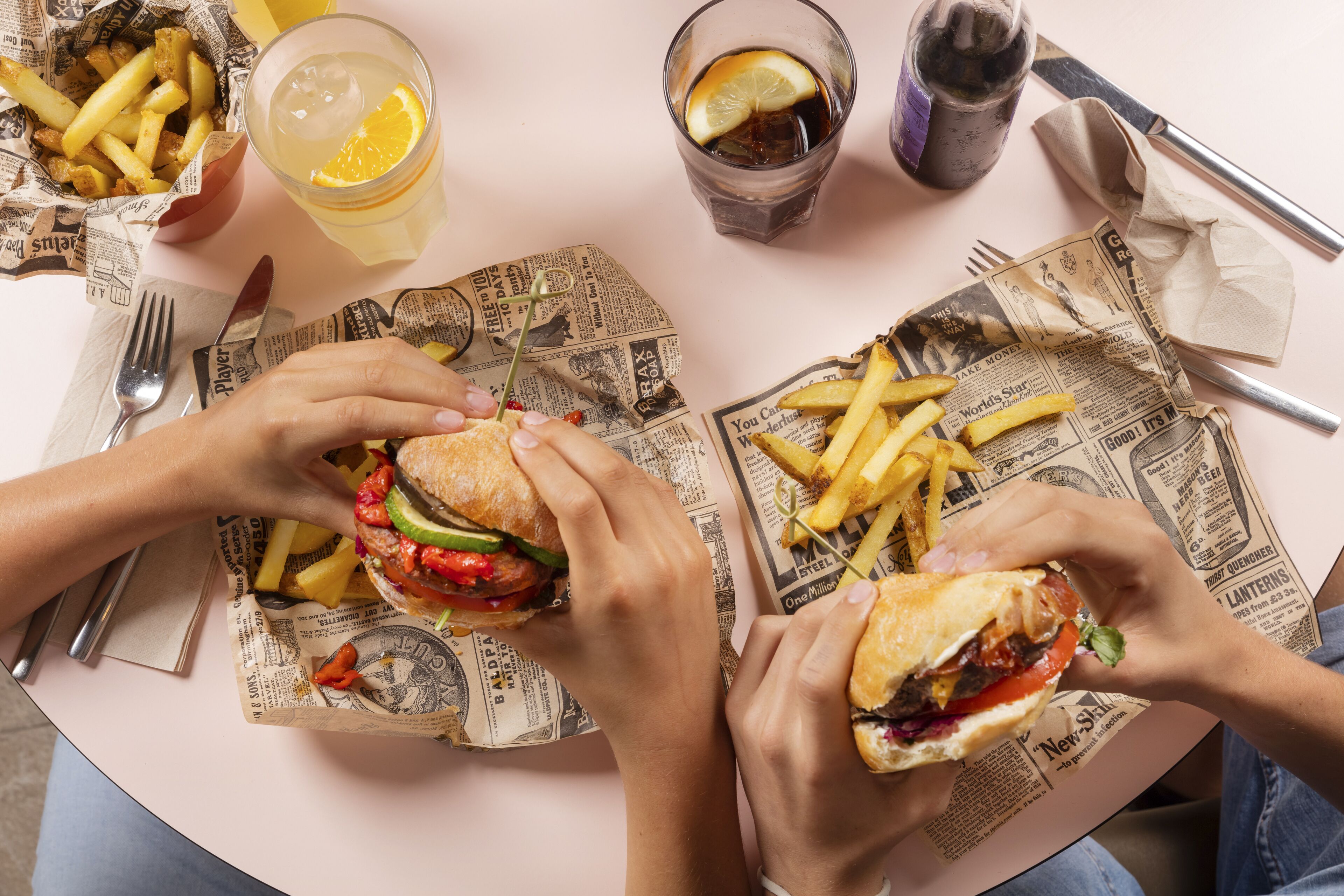 Una mesa con comida que incluye sándwiches y papas fritas, una bebida y cubiertos sobre un periódico.