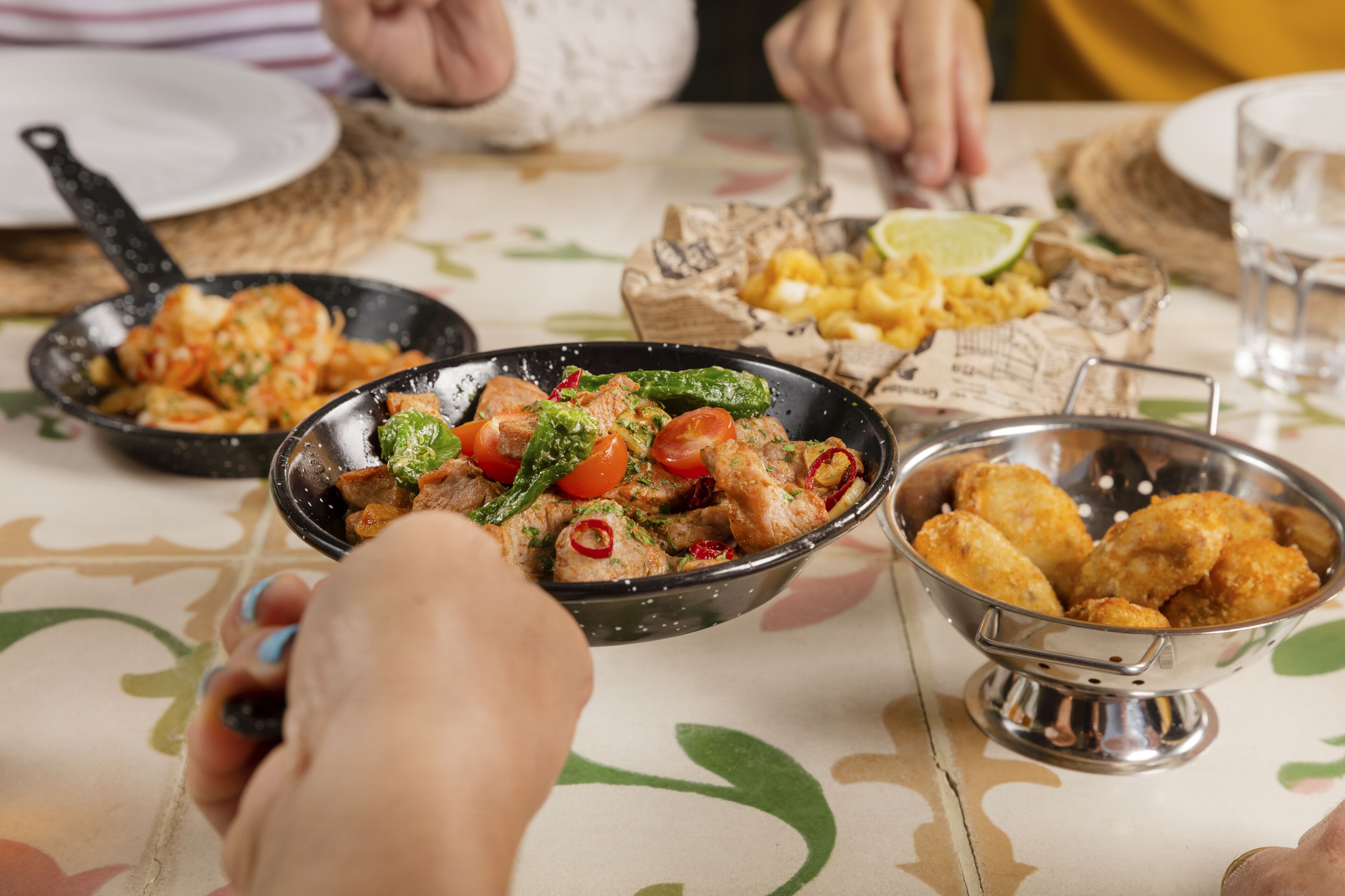 Una mesa de comedor con varios platos, incluyendo una sartén negra con comida, un bol metálico con alimentos fritos y un plato con un gajo de lima.