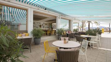 An outdoor patio area with round tables and chairs, potted plants, and a view of the sea.