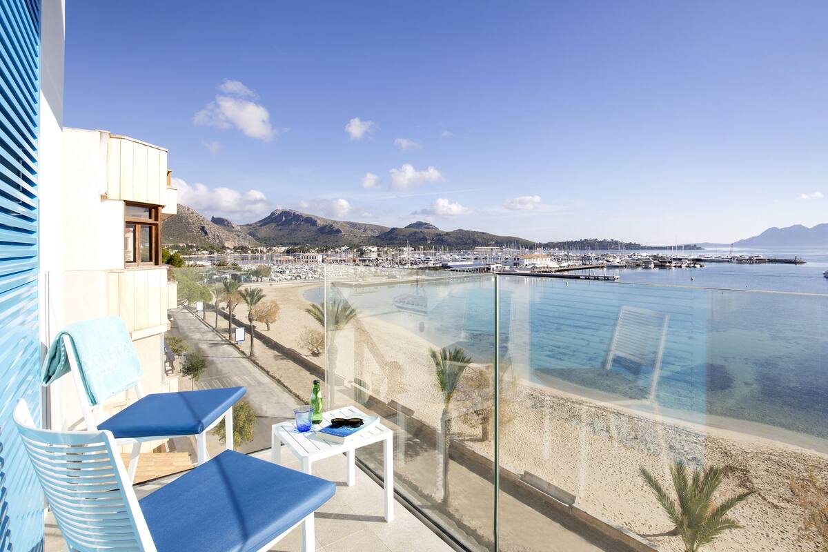A balcony with two blue lounge chairs, a small table with a bottle and glasses, and a view of a beach and marina.