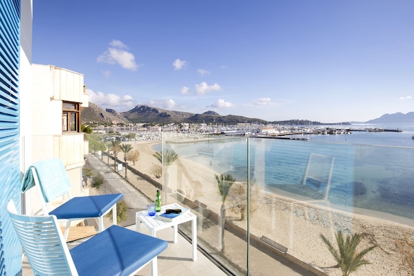 A balcony with two blue lounge chairs, a small table with a bottle and glasses, and a view of a beach and marina.