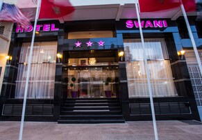 Una entrada de hotel con un llamativo letrero que dice 'HOTEL' y la marca 'SWANI'. Hay tres estrellas sobre la entrada, y el edificio tiene grandes ventanales con cortinas.