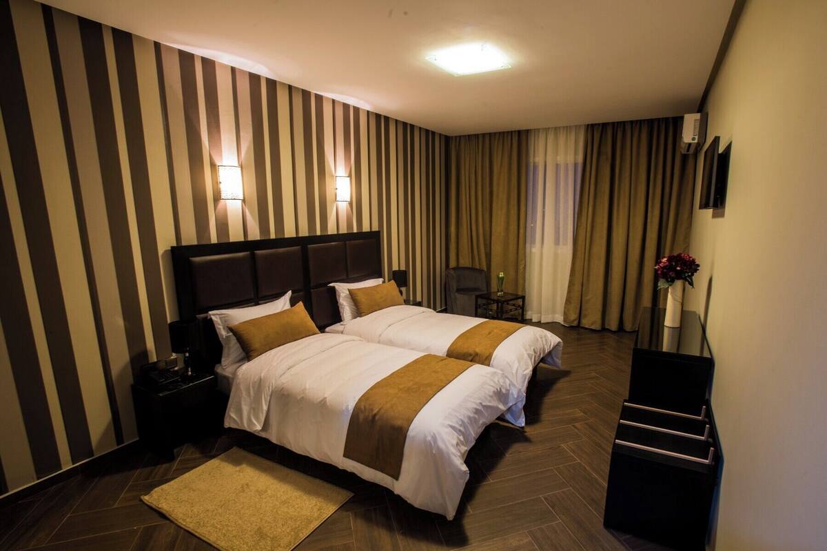 A hotel room with two beds, a desk, a chair, a small table with a vase, and a wall-mounted TV.