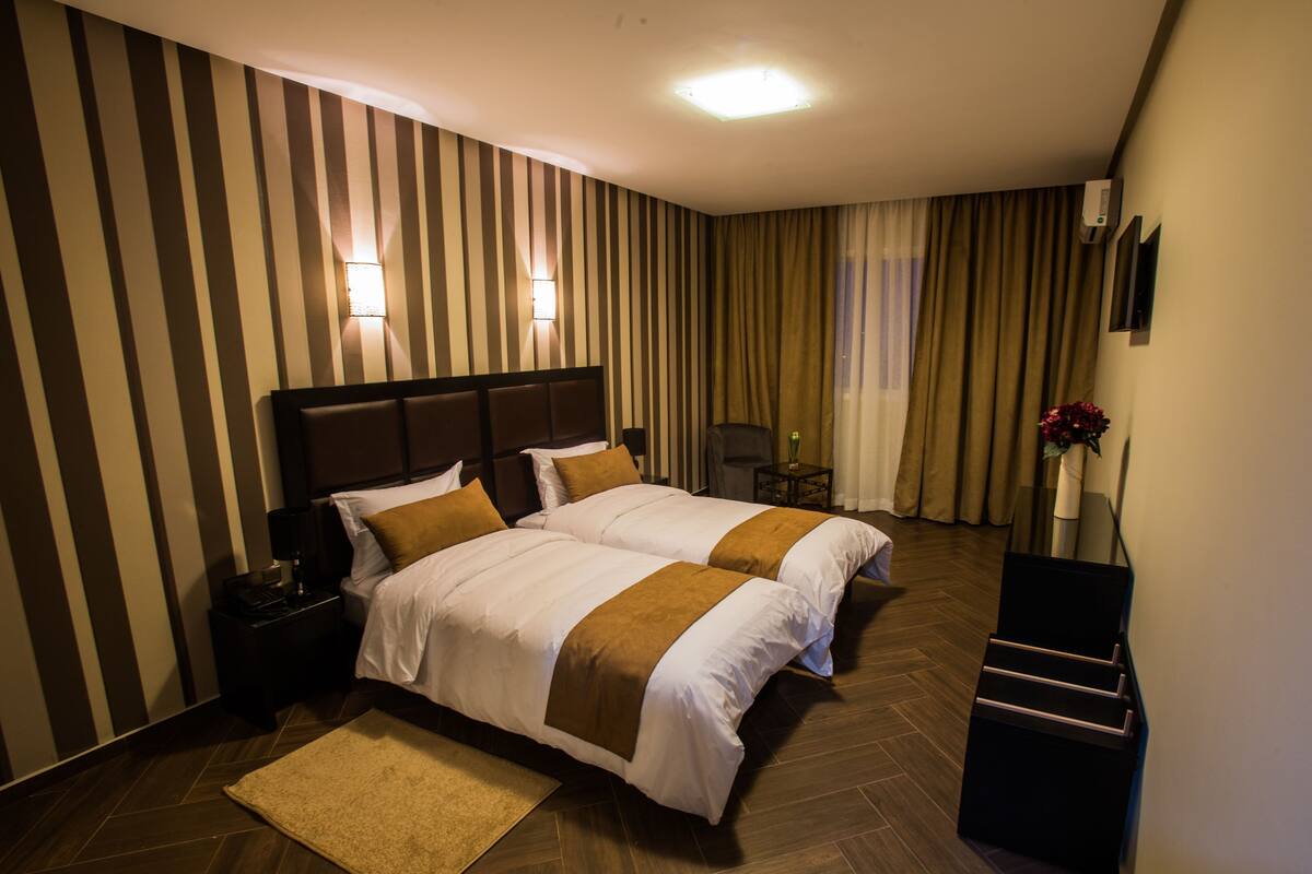 A hotel room with two beds, a nightstand, a chair, a small table with a vase, and a decorative plant.