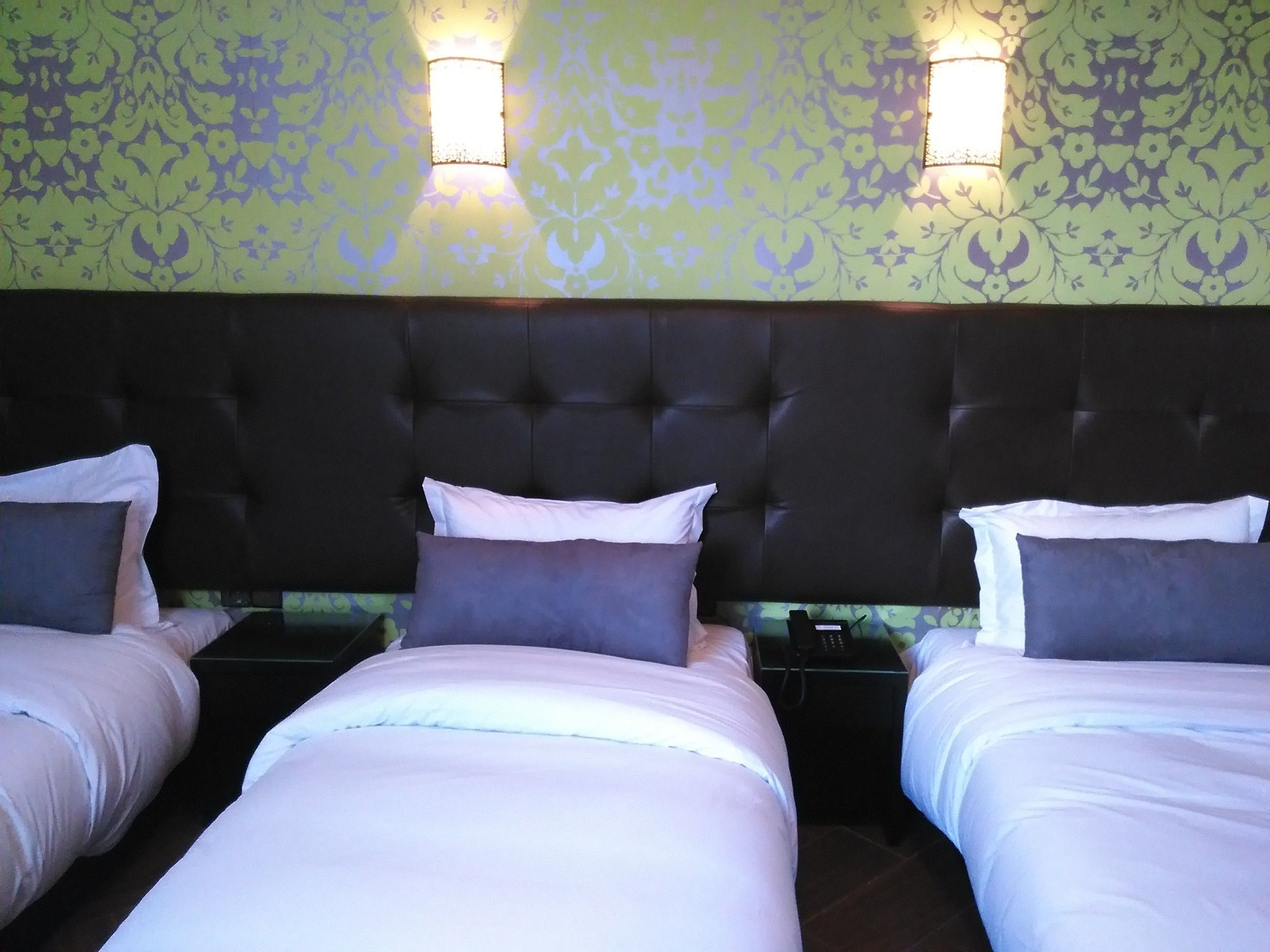 Habitación de hotel con tres camas, cada una con ropa de cama blanca y almohadas oscuras. Hay dos luces empotradas en el papel tapiz de diseño verde.