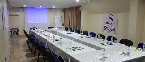 Un salón de conferencias con largas mesas, sillas, una pantalla de proyector y un cartel del 'Swan Hotel'.