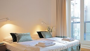 Habitación de hotel con dos camas, cada una con una almohada, una mesita de noche y una lámpara. Se ve una ventana con cortinas y vistas a edificios.
