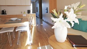 Una cocina moderna con armarios de madera, un comedor con una mesa de madera y sillas traslúcidas, y un florero con flores blancas sobre una mesita auxiliar.