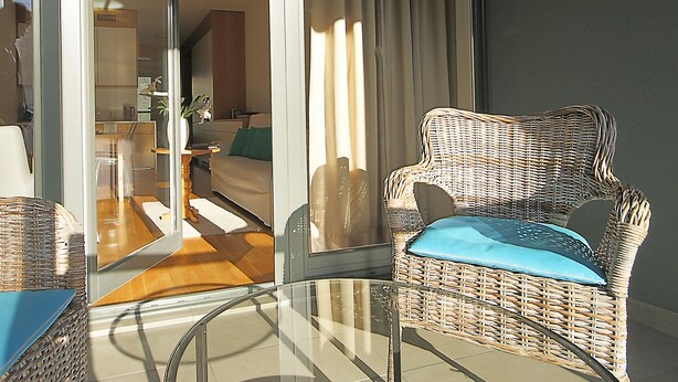 Una silla de mimbre con un cojín azul en una habitación soleada con una mesa de vidrio, un sofá y vistas de una cocina y sala de estar a través de una puerta de cristal.
