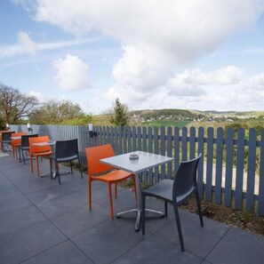 An outdoor patio with tables and chairs, a blue fence, and a view of a hilly landscape.