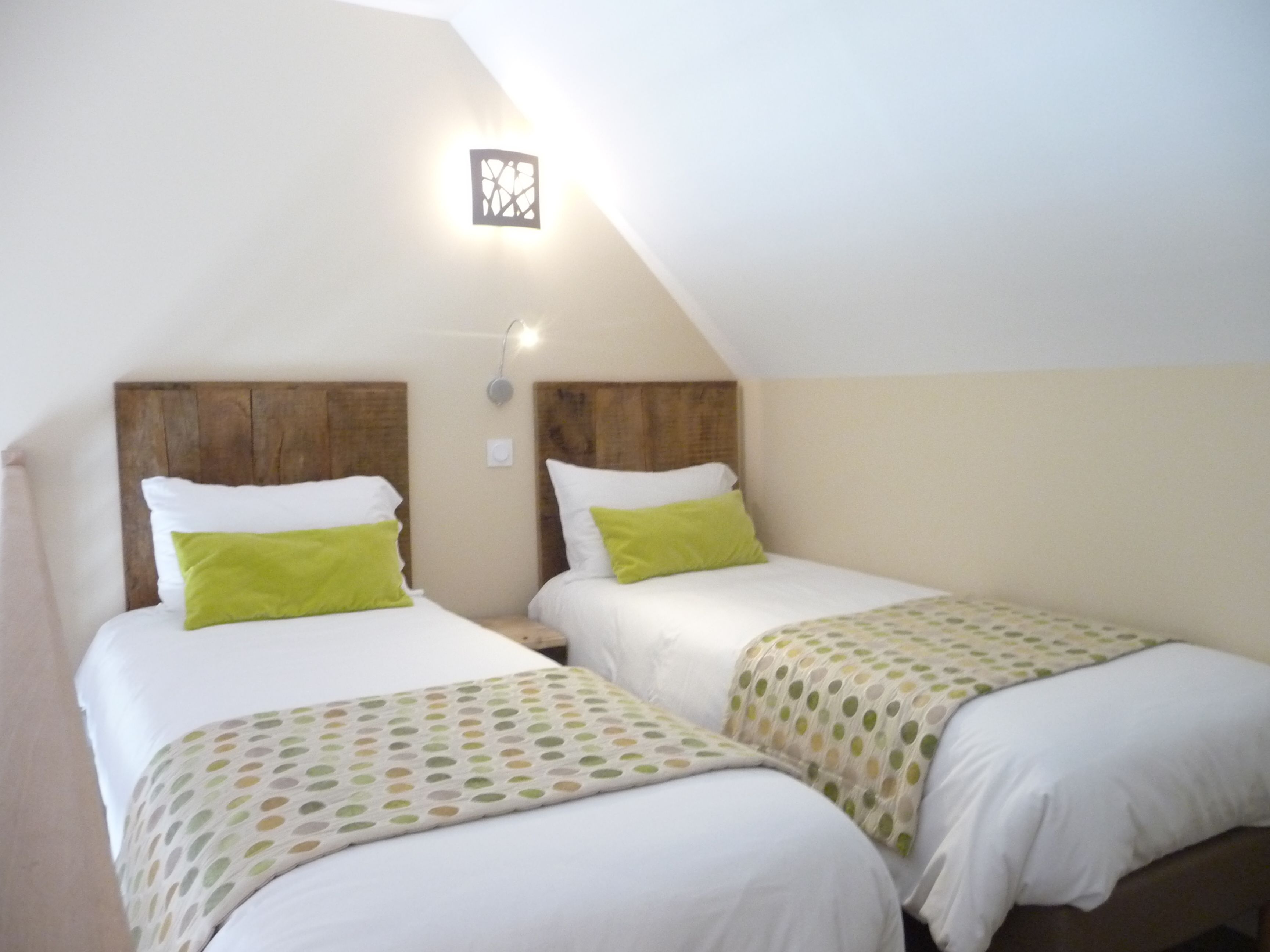 A room with two beds, each with white bedding and green pillows. There is a small wall-mounted light fixture above the beds.